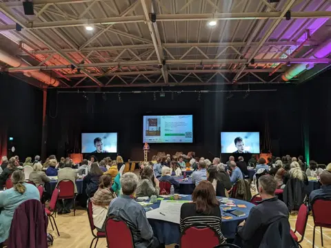 A large room with people seated around tables, the tables are covered with paper and pens. A screen at the far end shows a presentation.