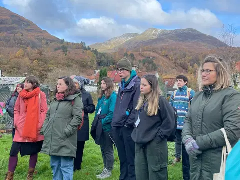 Scottish mountains in the background, some small buildings are visible. A group of people stand on grass in winter clothing watching a presentation.